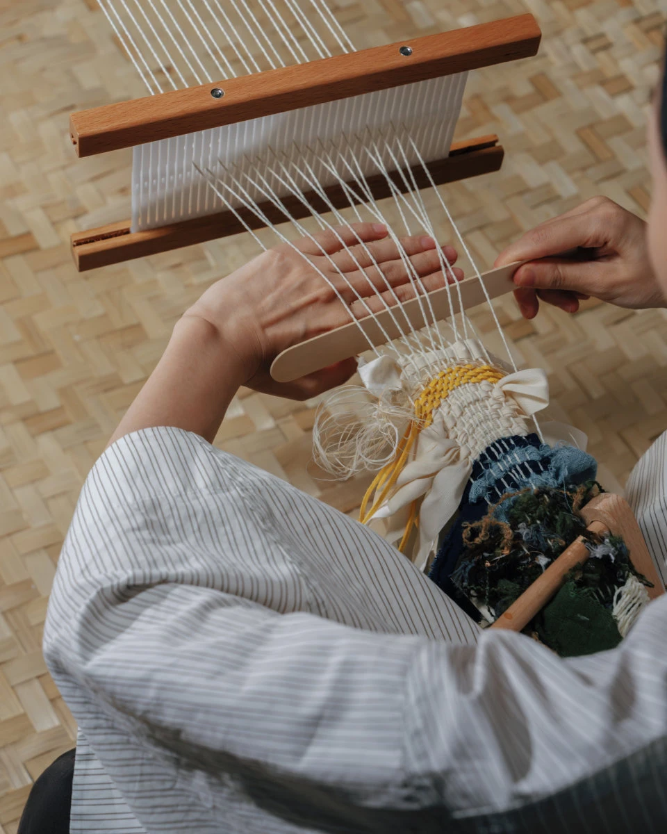 Hands working yarn and mixed materials on a small loom